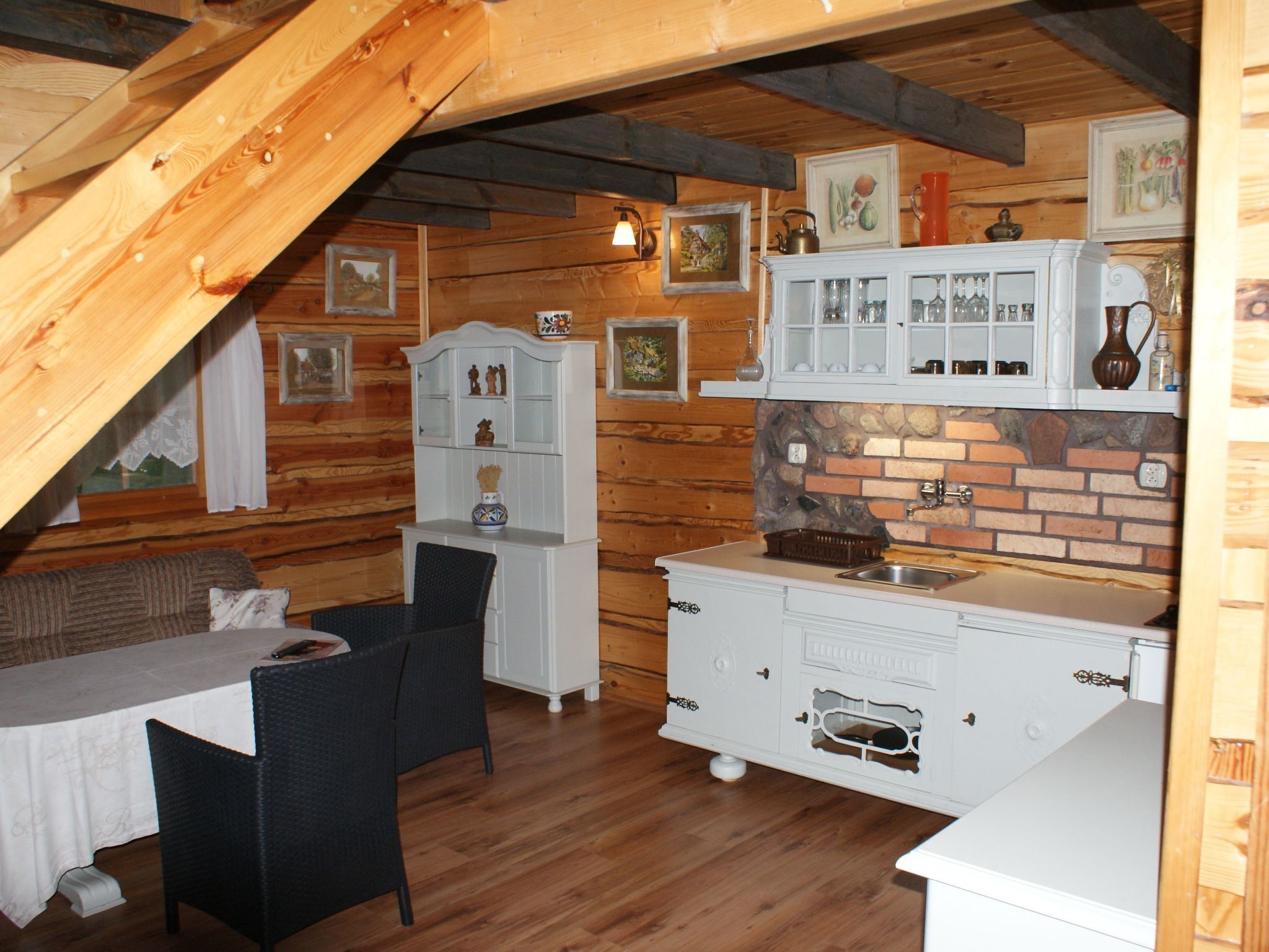 A cozy attic room with wooden beams, a dining table with chairs, a white cabinet with glass doors, and a brick backsplash.