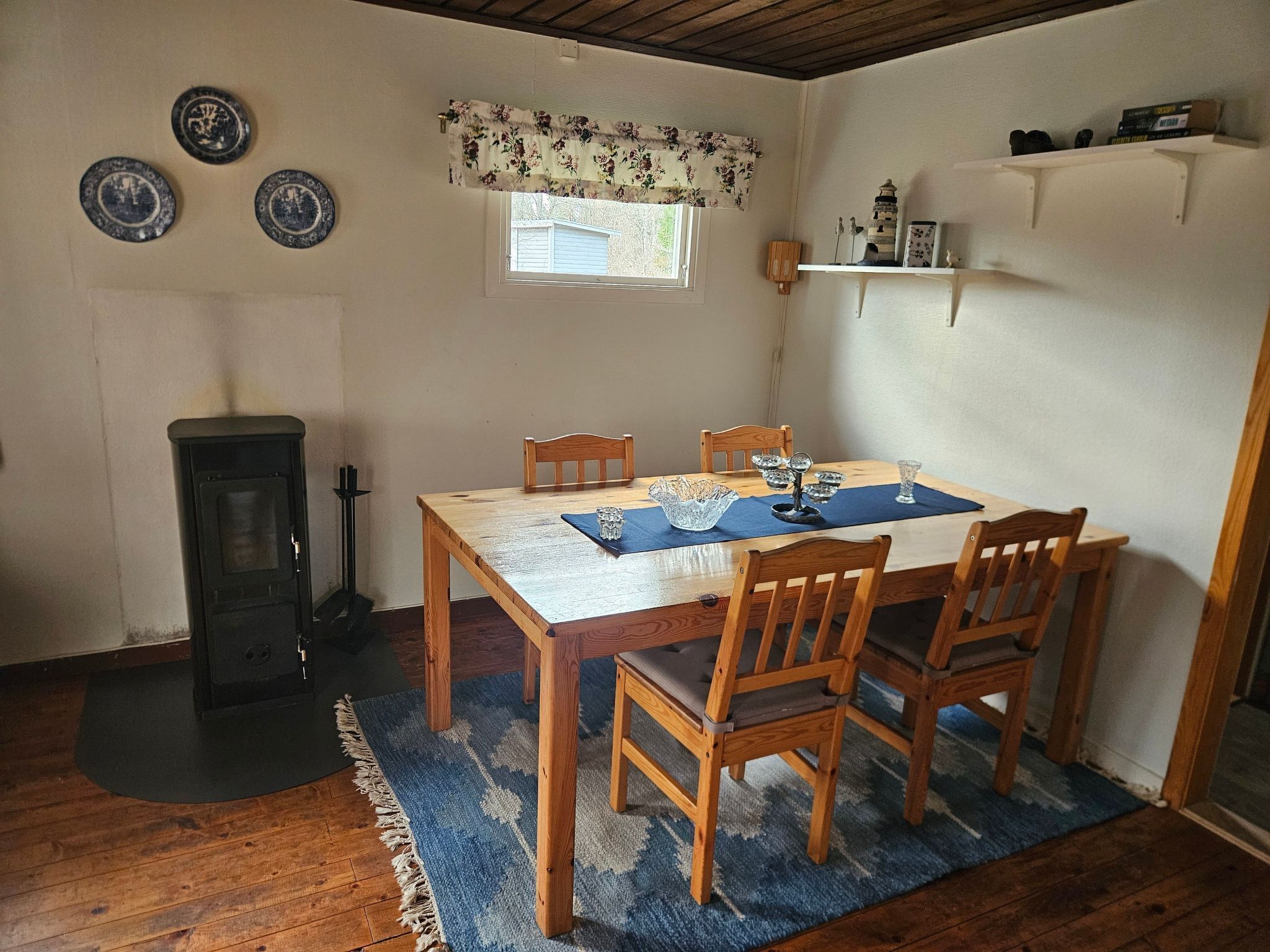 Ein Essbereich mit einem Holztisch, vier Stühlen, einem blauen Teppich und einem kleinen Fenster mit floralem Vorhang.