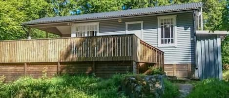A small, single-story cabin with a wooden deck and a covered porch, surrounded by lush greenery.