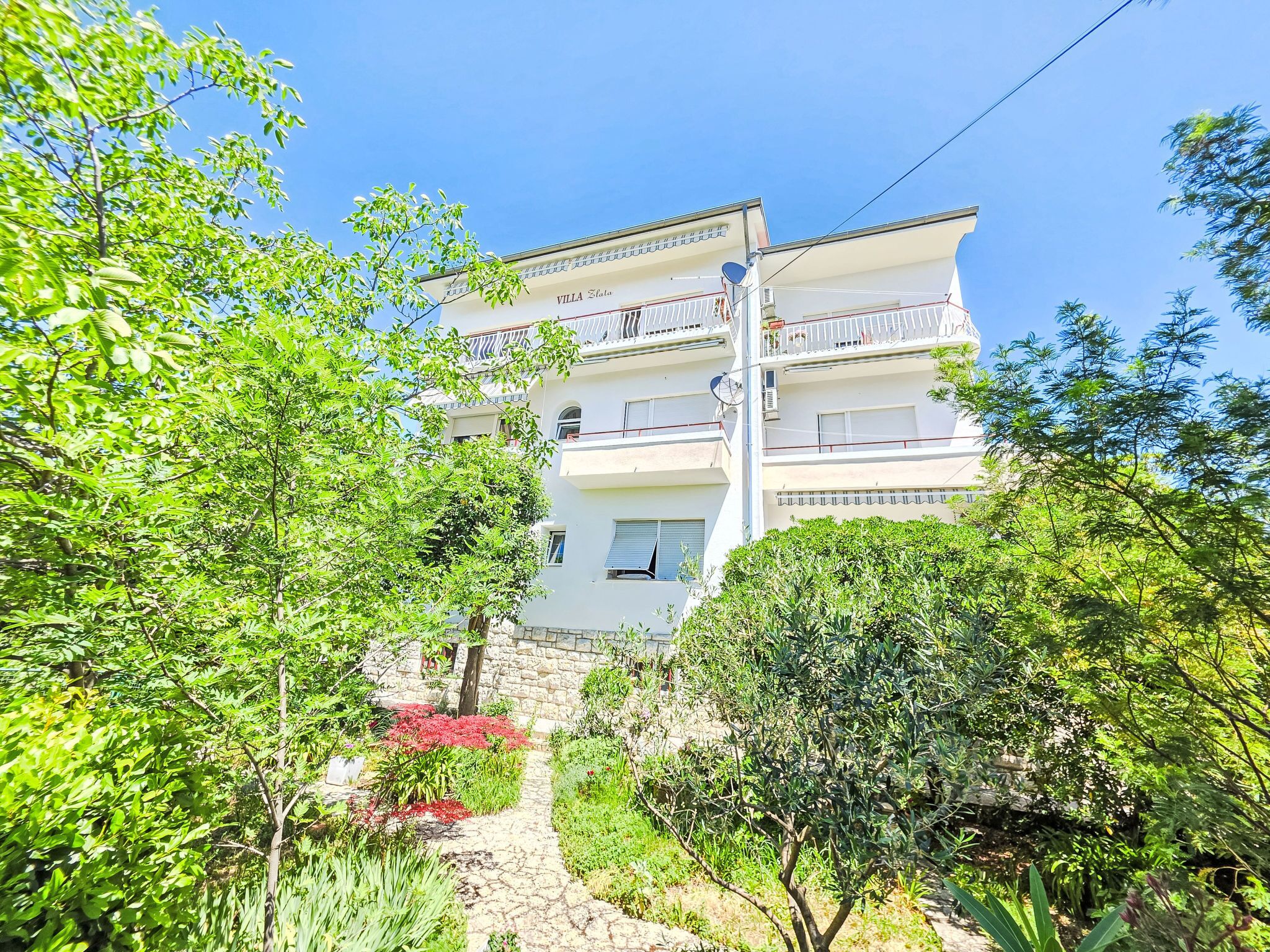 A multi-story building with balconies, surrounded by greenery and a stone pathway.
