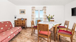 Ein Zimmer mit einem roten Sofa, einem Holzespeisetisch mit Stühlen, einem Holzschrank, einem an der Wand montierten Fernseher und einem Fenster mit Vorhängen.