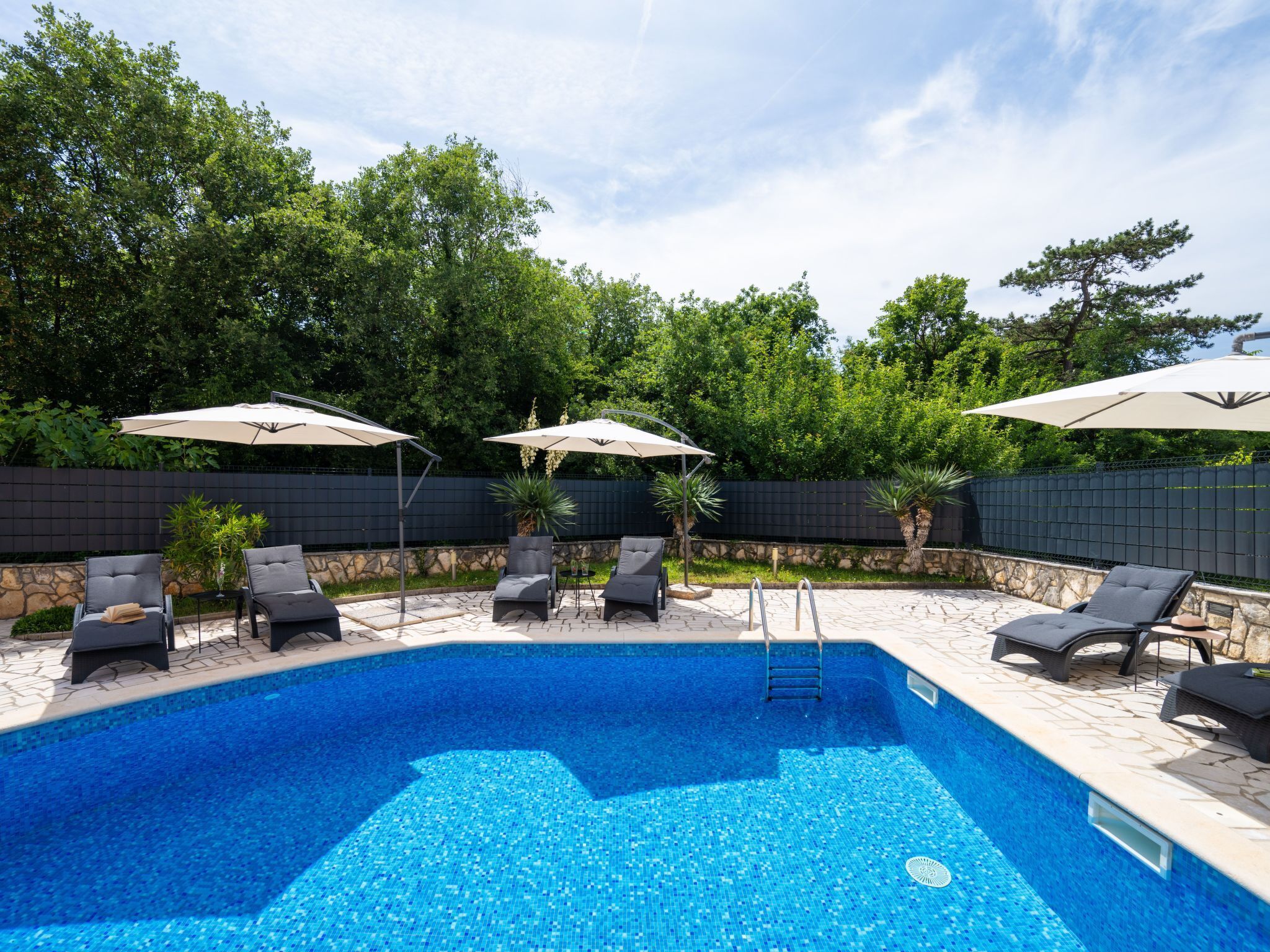 Una piscina con sillas de descanso y sombrillas, rodeada de un muro de piedra y vegetación.