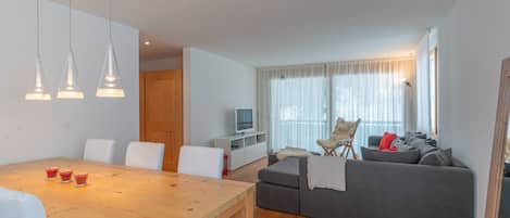 Uma sala de estar moderna com uma mesa de jantar de madeira, sofá cinzento e uma TV em um suporte branco.