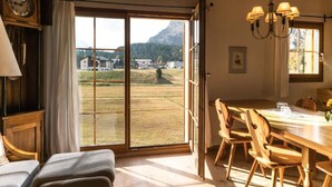 A room with a large sliding glass door leading to a view of a mountainous landscape. Inside, there is a wooden dining table with chairs, a wooden bench, and a wall-mounted clock. - Chesa Trais FluorsC3 by Interhome (Maloja)