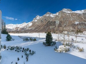 Schneebedeckte Landschaft mit Bergen im Hintergrund, blauem Himmel und vereinzelten Bäumen im Vordergrund.