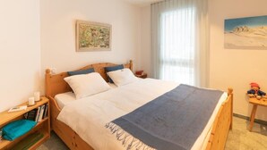 A bedroom with a wooden bed, a nightstand, a bookshelf, and a window with curtains. - Residenz Alpenrose34 by Interhome (Sils Maria)