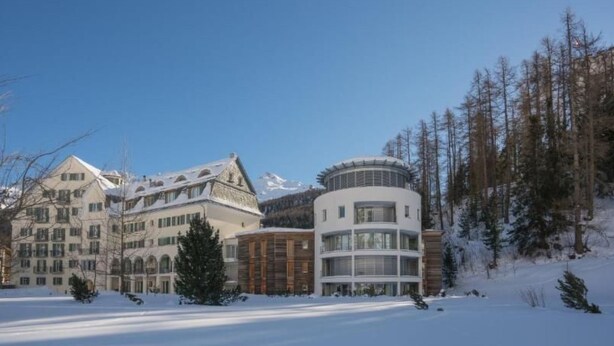 Ein schneebedecktes Hotel im traditionellen europäischen Baustil, mit mehreren Gebäuden, großen Fenstern und einer zentralen, runden Struktur.