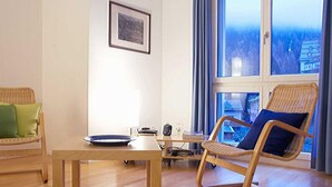 A modern living room with a wooden floor, a dining table, a wicker chair, and a large window with a view of a forested area. - Residenz Alpenrose22 by Interhome (Sils Maria)