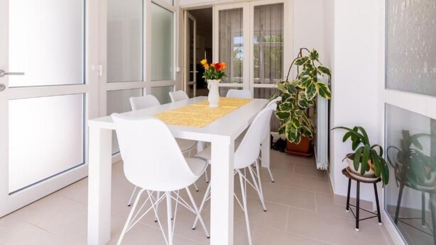 Un espace repas moderne avec une table et des chaises blanches, un vase rempli de fleurs et des plantes en pot.