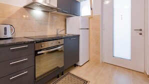 Une cuisine compacte équipée d’une cuisinière, d’un four et d’un petit réfrigérateur. Elle comprend une fenêtre avec des stores, une porte dotée d’un panneau en verre dépoli et un sol carrelé.