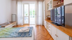 Une pièce lumineuse avec un plancher en bois, un lit recouvert d’une couvre-lit à motifs bleus et blancs, un téléviseur posé sur un meuble en bois et une grande fenêtre avec des rideaux.