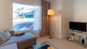 Ein modernes Hotelzimmer mit großem Fenster und Bergblick, einer Couch, einem Glastisch, einer Stehlampe und einem Fernseher auf einer Glassäule.