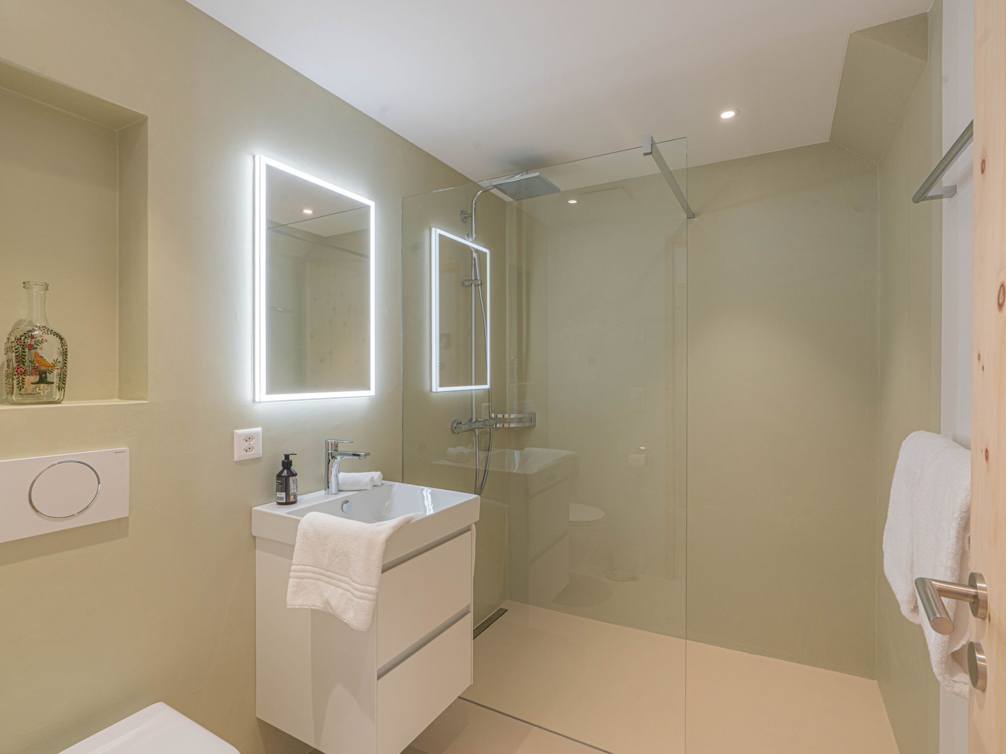 A modern bathroom with a glass-enclosed shower, a white sink with a mirror above it, and a bottle of shampoo on the counter.