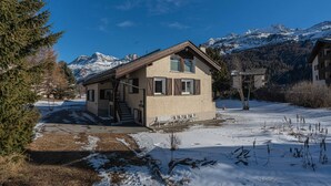 Ein Chalet mit geneigtem Dach, Holzfassade und einer Veranda. Umgeben ist es von verschneitem Gelände und Bergen im Hintergrund.
