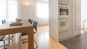 A modern kitchen with built-in appliances, a dining area with a wooden table and chairs, and large windows with sheer curtains. - Residenz Alpenrose15 by Interhome (Sils Maria)