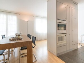 A modern kitchen with built-in appliances, a dining area with a wooden table and chairs, and large windows with sheer curtains. - Residenz Alpenrose15 by Interhome (Sils Maria)