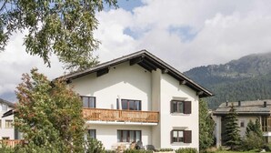 Un edificio de dos pisos con un balcón, rodeado de follaje y montañas al fondo.