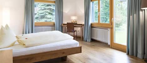Ein Schlafzimmer mit einem hölzernen Bett, einem Schreibtisch und Blick auf Grün durch große Fenster.