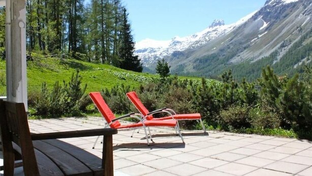 Un patio con dos sillones rojos y un banco de madera, con vistas a un paisaje montañoso con árboles y cielo despejado.