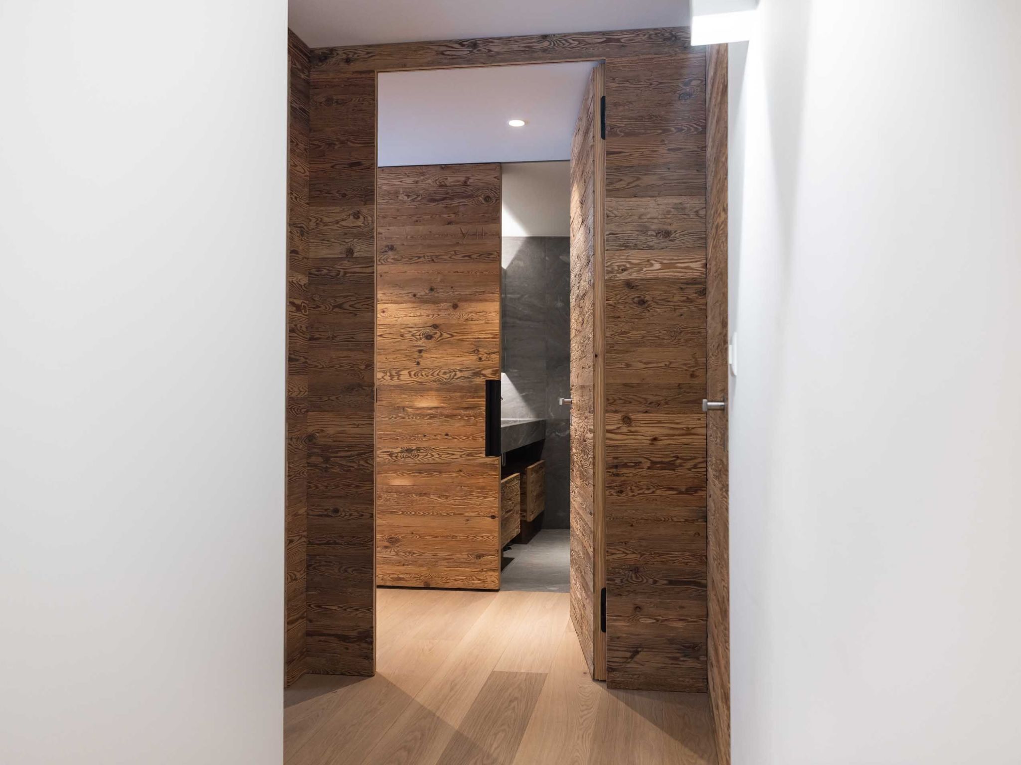 Un corridoio con rivestimenti in legno e una cucina moderna parzialmente visibile attraverso una porta aperta.