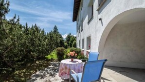 A patio with a table and chairs, surrounded by greenery and a building with an arched doorway. - Selvas 12 by Interhome (Sils Maria)