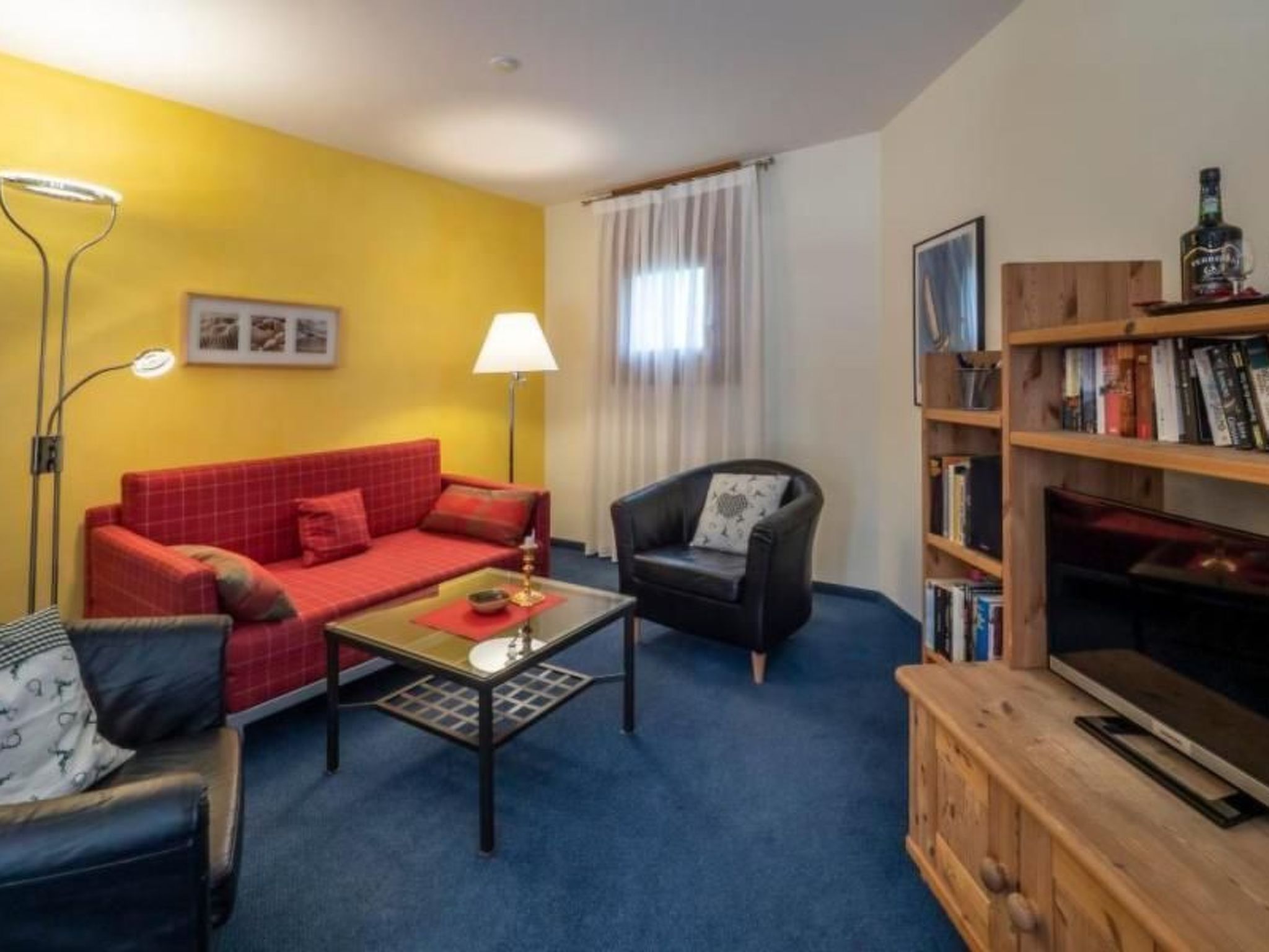 Ett rum med en röd soffa, en svart fåtölj, ett soffbord, en lampa, en bokhylla och en tv.