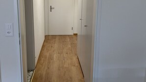 Un couloir au sol en bois et aux murs blancs, qui mène à une porte au fond.