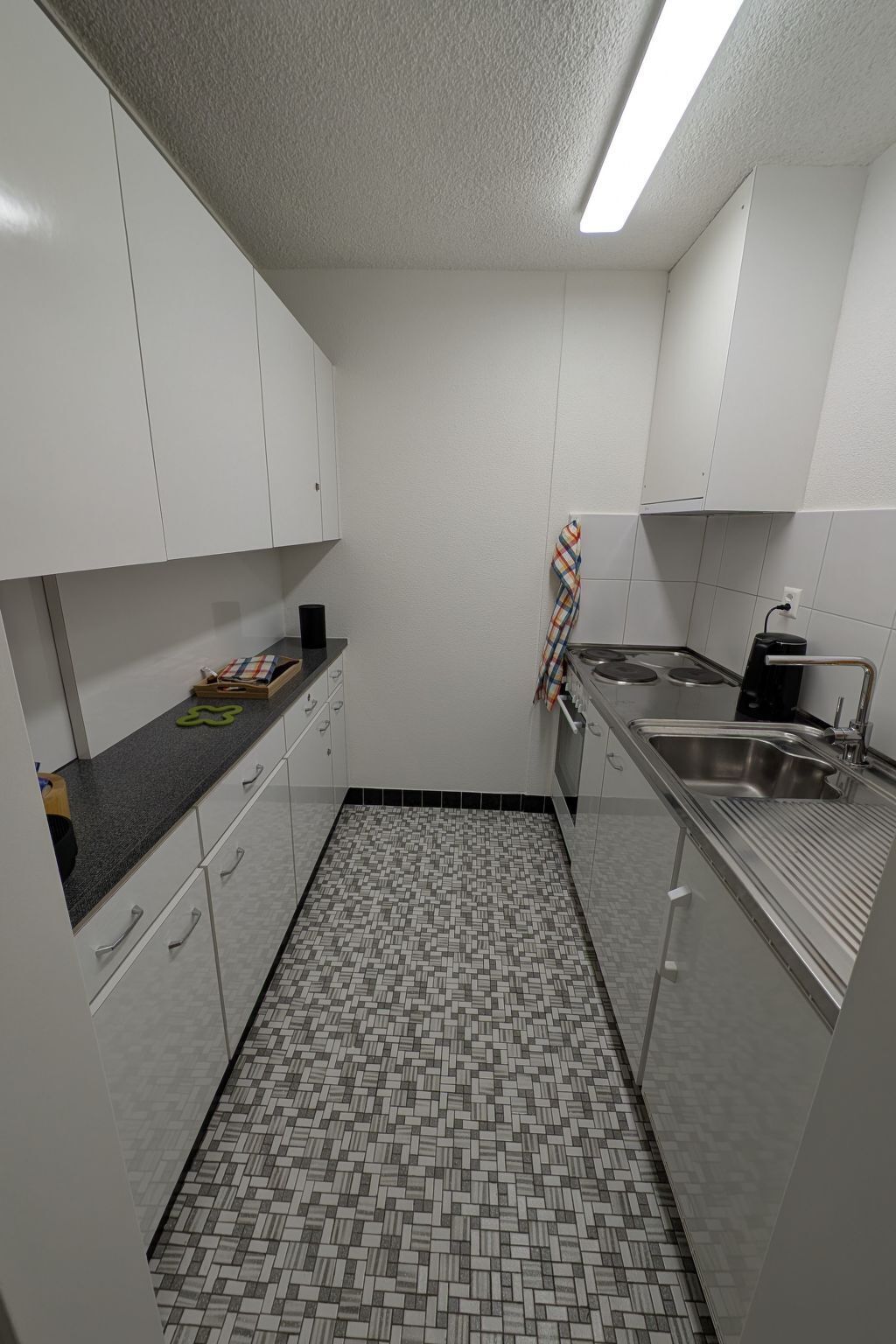 Una cocina compacta con gabinetes blancos, piso de baldosas y electrodomésticos de acero inoxidable.