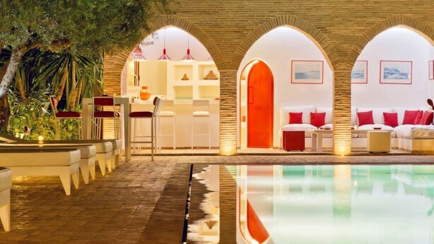 Un espace aménagé au bord de la piscine offrant une vue dégagée sur celle-ci, un coin salon avec des coussins rouges et un comptoir d’accueil doté d’une porte d’un orange vif.