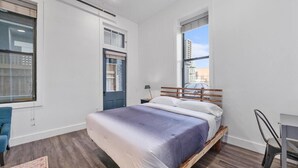 Un dormitorio moderno con una cama grande, un escritorio y una silla. Hay ventanas que ofrecen luz natural y vistas de la ciudad.