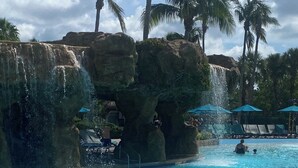 Una piscina azul cristalina con una cascada, rodeada de palmeras y sillas de descanso.