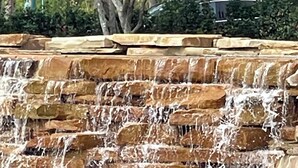 Una cascada se precipita sobre escalones de piedra escalonados hacia una piscina que refleja el entorno, con una garza de pie en el agua.