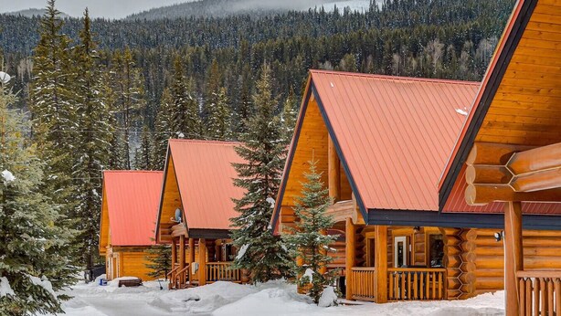 A row of log cabins with red roofs, surrounded by snow-covered ground and evergreen trees, set against a backdrop of a forested mountain.