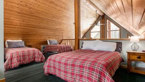 A cozy attic bedroom with wooden walls and sloped ceilings, featuring two beds with plaid bedding and a small bedside table with a lamp.
