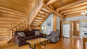 A log cabin interior with a wooden staircase, a brown leather sofa, a patterned armchair, and a wooden dining table.