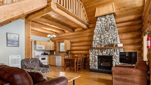A log cabin interior with a stone fireplace, wooden dining table, and leather sofas.