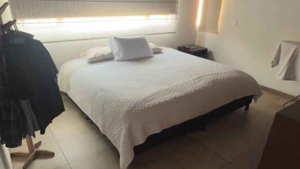 Une chambre à coucher comprenant un lit, une table de chevet, une fenêtre avec des stores et un porte-manteau.