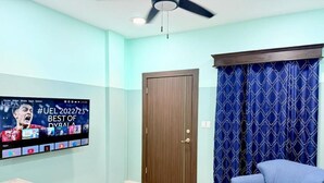 Una habitación con un televisor montado en la pared mostrando la interfaz de una aplicación de música, una puerta de madera, una cortina azul estampada y un sillón azul.