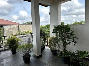 Un balcón cubierto con plantas en macetas, pilares de hormigón y vista a un edificio de techo rojo al fondo.