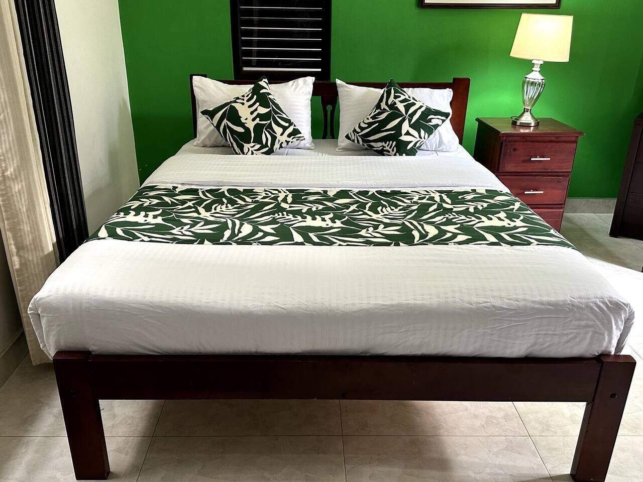Un letto con copriletto bianco e verde fantasia, una testata in legno e un comodino con una lampada.
