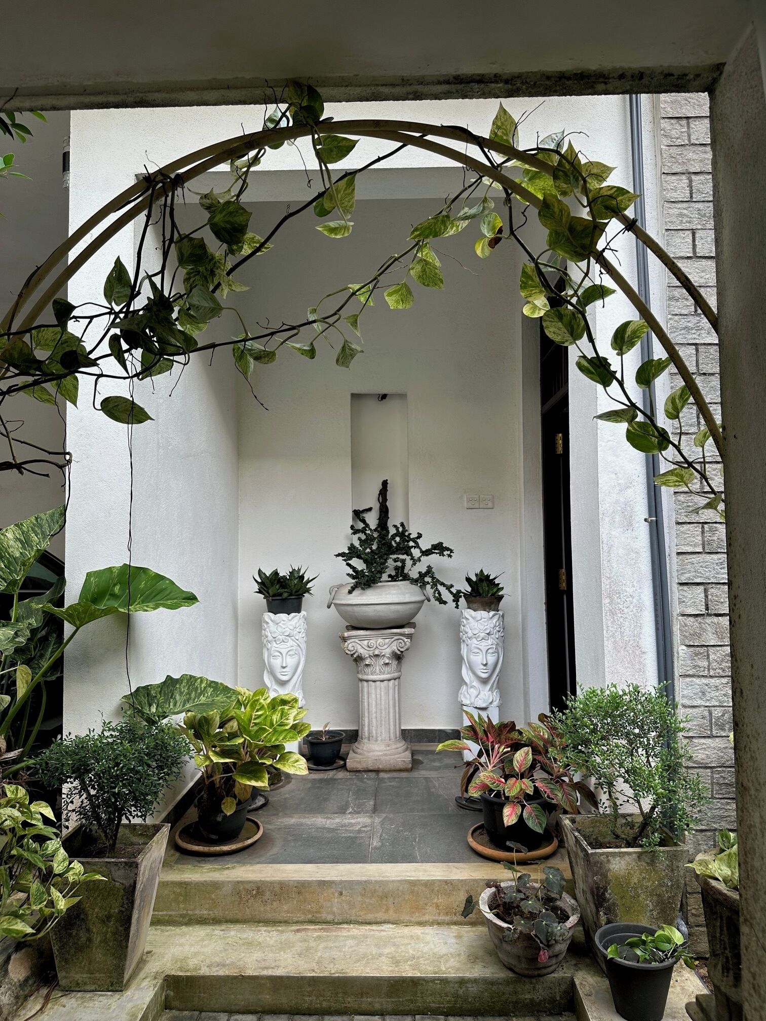 Een stenen trap die leidt naar een deur, omringd door verschillende plantenpotten en een decoratieve boog.