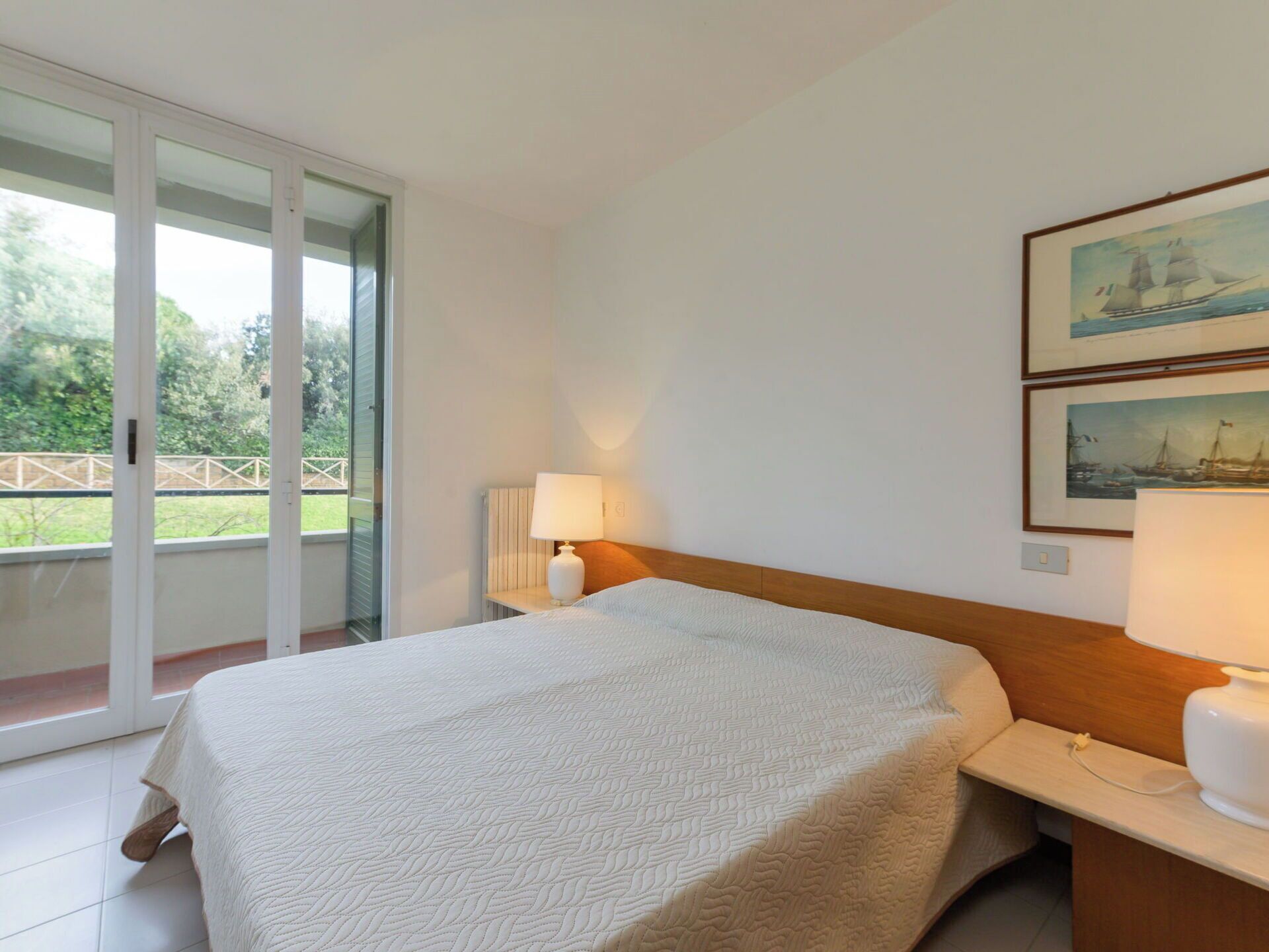 A hotel room with a bed, a nightstand with a lamp, a sliding glass door to a balcony, and two framed pictures on the wall.