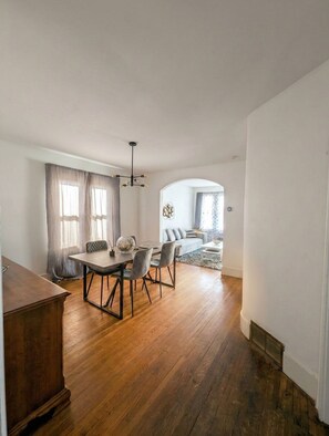 A spacious room with a wooden floor, a dining table with chairs, a sofa, and a large window with curtains.