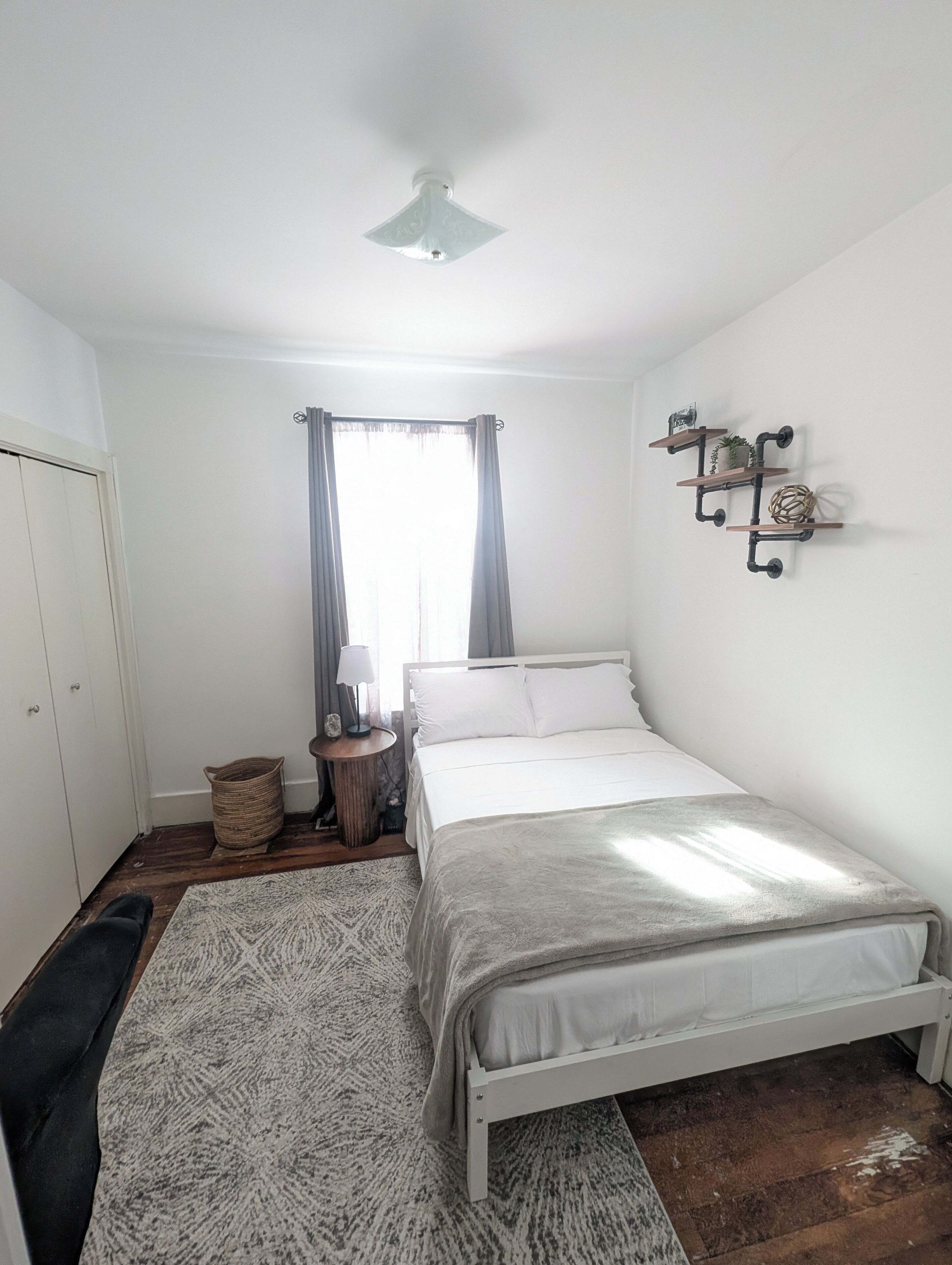 Une chambre à coucher comprenant un lit, une table de chevet, une lampe, une étagère murale décorative, un ventilateur de plafond et une fenêtre avec des rideaux.