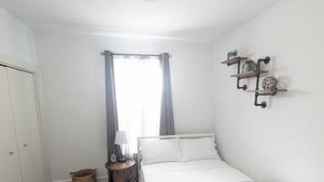 Une chambre à coucher comprenant un lit, une table de chevet, une lampe, une étagère murale décorative, un ventilateur de plafond et une fenêtre avec des rideaux.