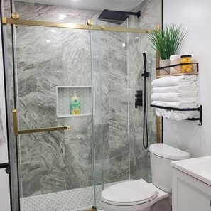 A bathroom with a glass-enclosed shower, a toilet, a shelf with toiletries, and a towel rack.
