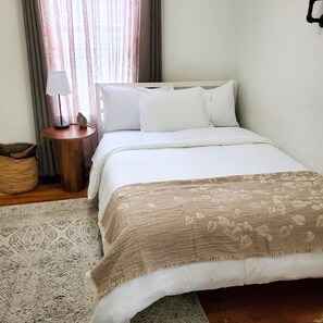 A neatly made bed with white linens and a beige blanket, a bedside table with a lamp, a woven basket, and a window with curtains.