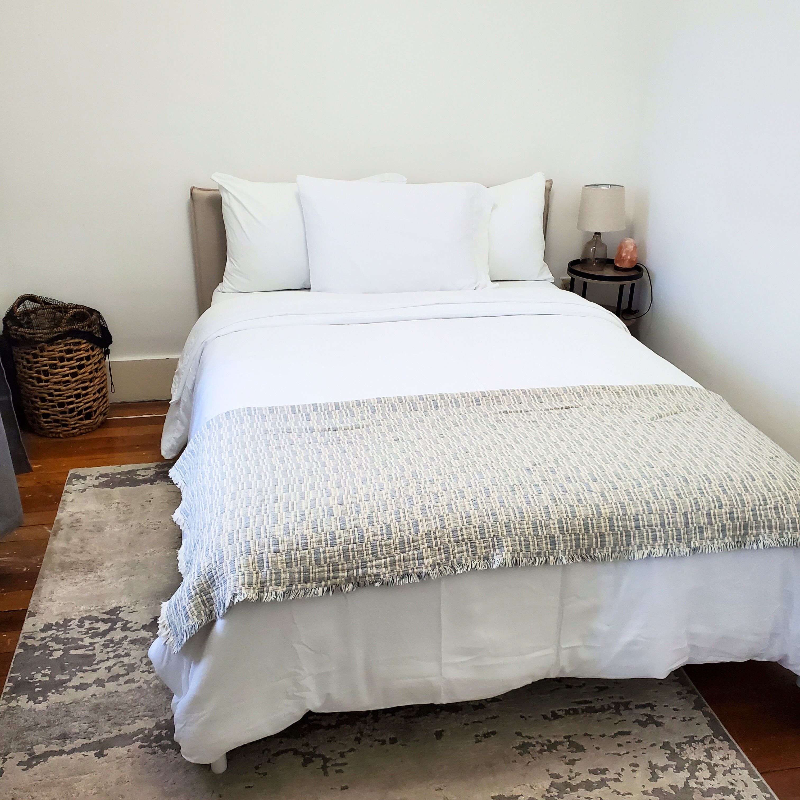 A neatly made bed with white bedding, a patterned throw blanket, and a small bedside table with a lamp and a decorative item.