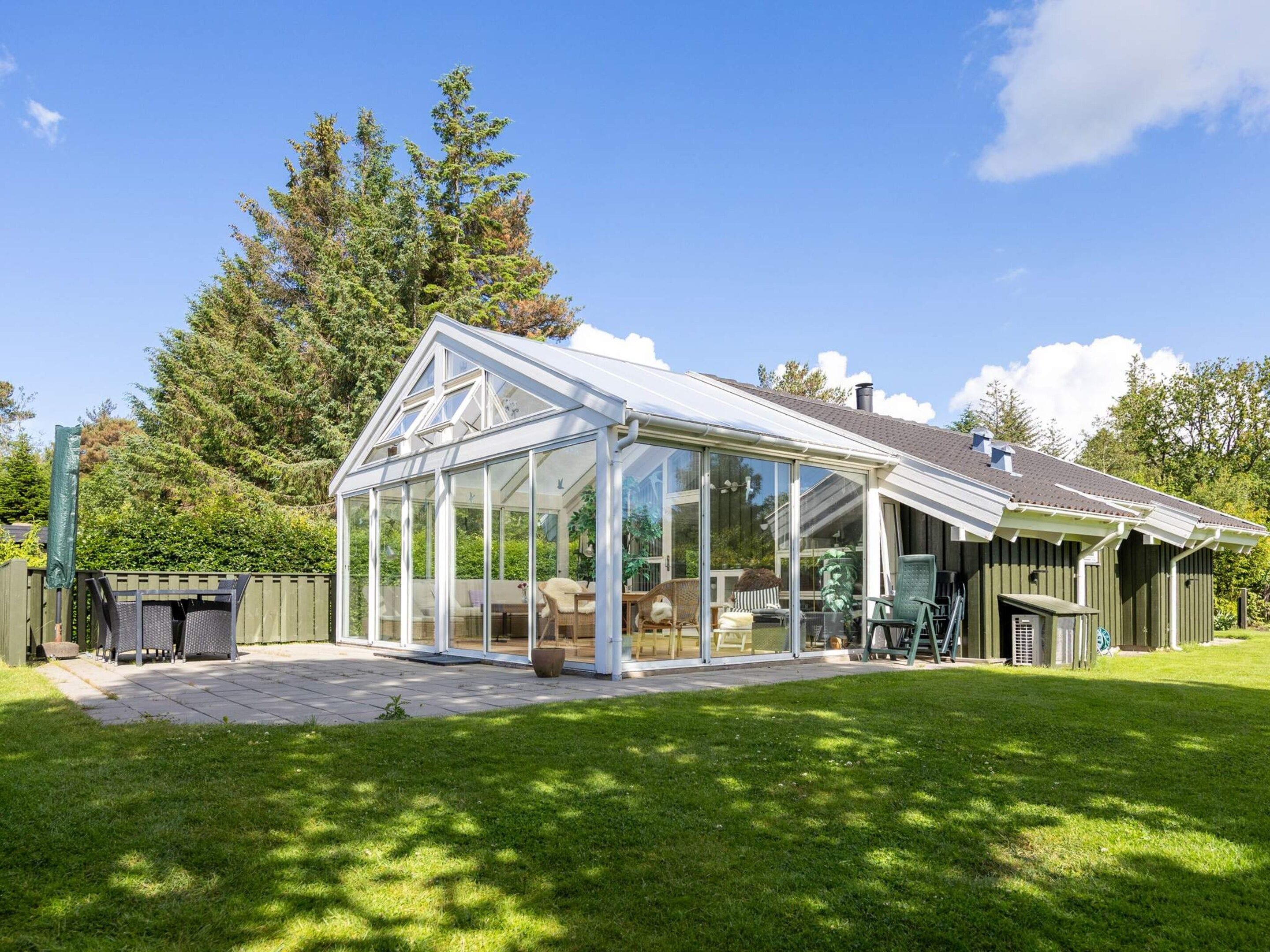 A glass conservatory attached to a house with a sloped roof, surrounded by a well-maintained lawn and trees.