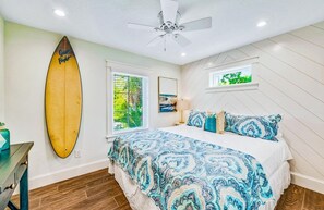 A bedroom with a bed, a ceiling fan, a surfboard, and a window with a view of greenery. - Luxury House w/ Heated Pool & Spa, Walk to Beach (Siesta Key)